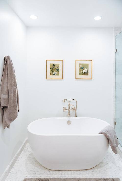 Bathroom design displaying honed white marble grid floor tiles fitted with an oval bathtub joined with a vintage handheld tub filler against a wall showcasing botanical art in gold frames.