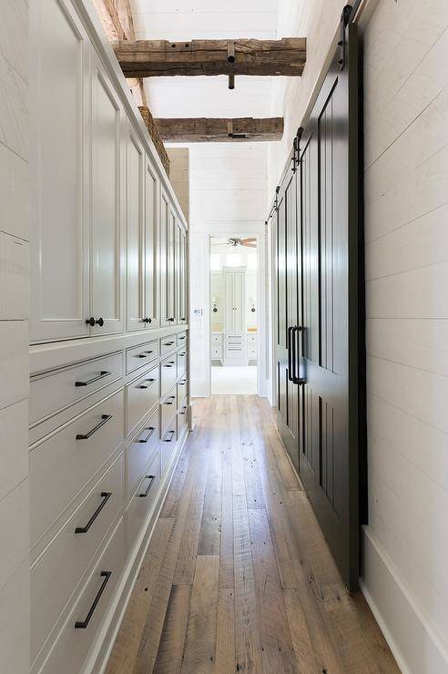A beautifully fitted bathroom hallways boasts gray built-in cabinets donning oil rubbed bronze pulls and positioned facing a black barn door on rails and beneath rustic wood ceiling beams fixed between white shiplap walls.