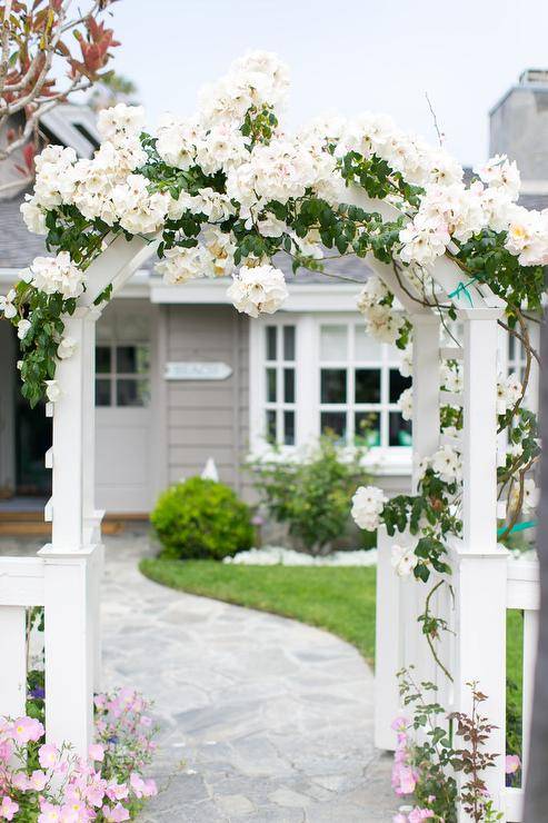 A winding stone pathway flanked by a white picket fence leads through a flower covered front gate arbor.