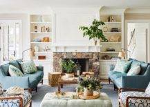 White built-in shelves mounted over white cabinets flank a fireplace boasting a stone surround a white mantel. A white canvas art piece hangs over the mantel and faces a brown tray coffee table placed between facing blue striped sofas placed on a blue striped rug.