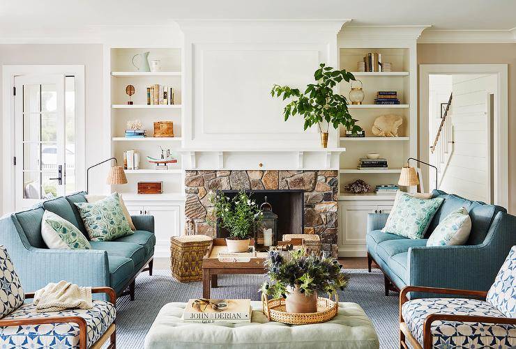 White built-in shelves mounted over white cabinets flank a fireplace boasting a stone surround a white mantel. A white canvas art piece hangs over the mantel and faces a brown tray coffee table placed between facing blue striped sofas placed on a blue striped rug.