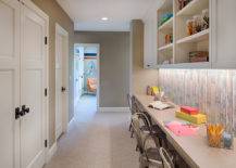 Hallway homework station with built-in upper cabinets with open shelving over a weathered plank effect backsplash alongside a wall to wall taupe counter lined with Tolix chairs atop beige wall to wall carpeting.