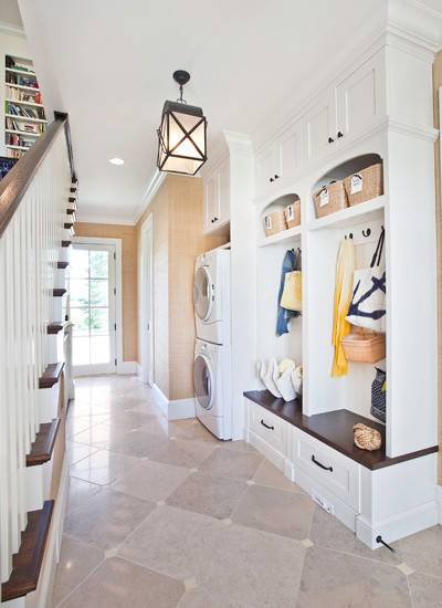 Beautiful hall and stairway with limestone tiled floors laid in a diamond pattern and grasscloth wallpaper. The mudroom area features built-in arched cubbies with bench and drawer storage. Overhead cubbies hold wicker baskets with closed cabinetry above. A stackable front loading washer and dryer sit beside the mudroom area inside a built-in nook. A unique lantern style pendant hangs over the space.