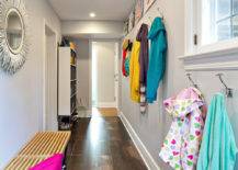 Family friendly hallway mudroom with oversized tiled floors and gray walls. Below the Horchow Janice Minor White "Porcupine Quill" Mirror stands a contemporary wooden bench. At the end of the hallway stands open cubby storage for the families outdoor wear. In the foreground coat hooks line the wall with a cable picture hanging system holding colorful kids artwork above.