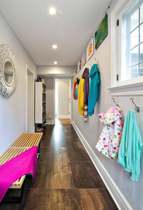 Family friendly hallway mudroom with oversized tiled floors and gray walls. Below the Horchow Janice Minor White "Porcupine Quill" Mirror stands a contemporary wooden bench. At the end of the hallway stands open cubby storage for the families outdoor wear. In the foreground coat hooks line the wall with a cable picture hanging system holding colorful kids artwork above.