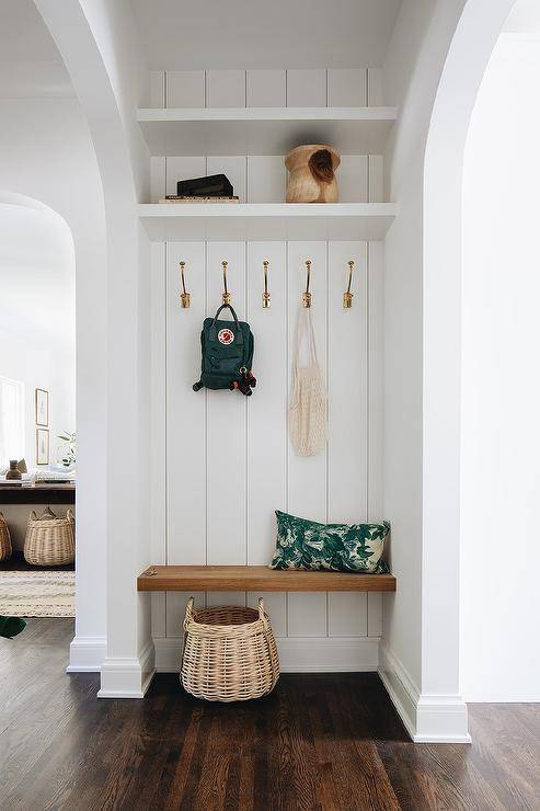 A window floating bench is fixed in an entry nook against shiplap trim holding brass coat hooks under stacked white shelves.