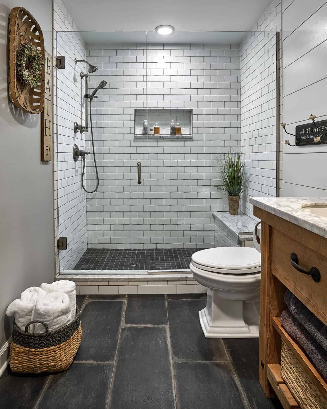 white subway tile farmhouse basement bathroom with wood vanity toilet grey slate floor tobacco basket hanging on wall basket of rolled towels