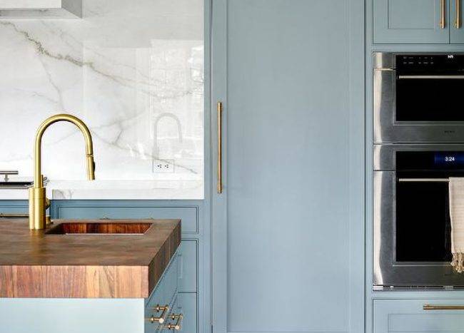 A butcher block countertop accents a blue kitchen island finished with a sink and a polished nickel gooseneck faucet. A blue wood paneled fridge door with a brass handle is fixed beneath blue cabinets and beside a wall oven fixed under a microwave.