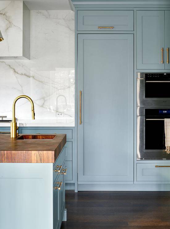 A butcher block countertop accents a blue kitchen island finished with a sink and a polished nickel gooseneck faucet. A blue wood paneled fridge door with a brass handle is fixed beneath blue cabinets and beside a wall oven fixed under a microwave.