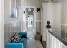 A second floor landing is filled with a blue velvet tufted bench lining a gray beadboard wall tucked under side by side blue art pieces. A gray trellis runner leads to a white freestanding linen cabinet adorned with brass door handles.