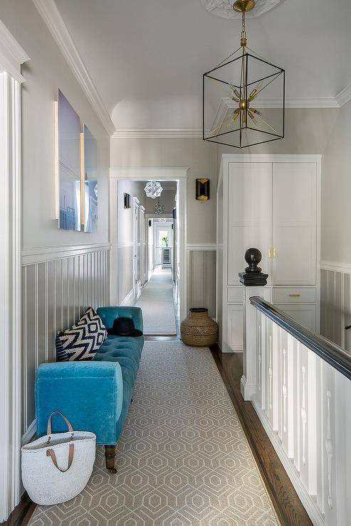A second floor landing is filled with a blue velvet tufted bench lining a gray beadboard wall tucked under side by side blue art pieces. A gray trellis runner leads to a white freestanding linen cabinet adorned with brass door handles.
