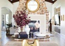 Large foyer features a walnut and brass table in the center atop a taupe rug and a round gold mirror mounted over a double sided stone fireplace.