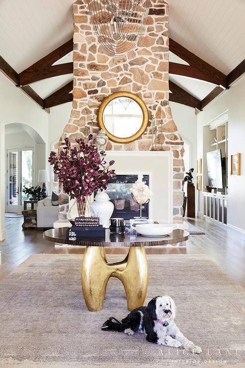 Large foyer features a walnut and brass table in the center atop a taupe rug and a round gold mirror mounted over a double sided stone fireplace.