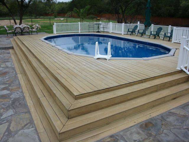 large wood deck built around a rectangle above ground pool patio chairs and white fence