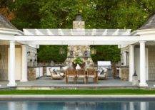 A teak dining table surrounded by teak dining chairs sits on a patio in front of an in-ground swimming pool. The patio is finished with a stone outdoor kitchen wrapping around to a gray stone fireplace.