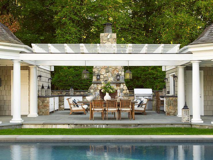 A teak dining table surrounded by teak dining chairs sits on a patio in front of an in-ground swimming pool. The patio is finished with a stone outdoor kitchen wrapping around to a gray stone fireplace.