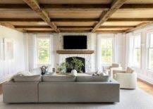 Living room features a rustic plank coffered ceiling over a white stone fireplace, ivory teddy boucle chairs with a charcoal gray corner sectional and a round beige wooden coffee table atop a gray rug