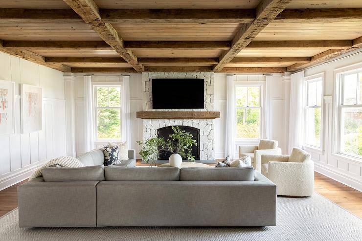 Living room features a rustic plank coffered ceiling over a white stone fireplace, ivory teddy boucle chairs with a charcoal gray corner sectional and a round beige wooden coffee table atop a gray rug