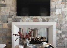 Living room features a tv mounted over a white beveled double sided mantel on gray and gold stone fireplace and a square wooden coffee table atop a gray rug.