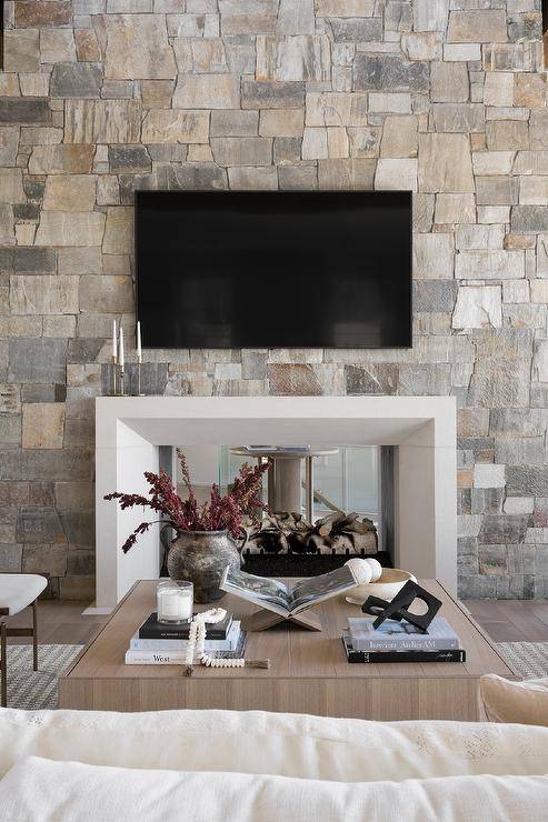 Living room features a tv mounted over a white beveled double sided mantel on gray and gold stone fireplace and a square wooden coffee table atop a gray rug.