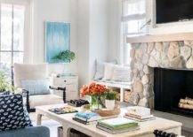 Welcoming living room features a TV fixed in a shiplap niche over a rustic gray stone fireplace. A light stained wooden coffee table sits on a blue rug in front of a blue sofa.