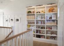 Second floor landing hall features built in bookcase illuminated with satin nickel picture lights beside walls clad in board and batten lined with black and white family photos.