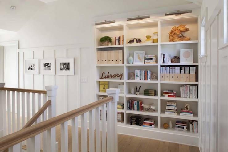 Second floor landing hall features built in bookcase illuminated with satin nickel picture lights beside walls clad in board and batten lined with black and white family photos.