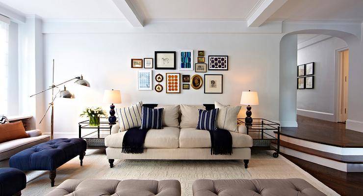 Living room outfitted with large cream silk rug, English rolled arm seating, linen & down pillows, and a collage of modern and vintage artwork, custom built-ins with blue backs. On the far left wall, two African petrified animal heads hang to add interest to this corner of the room. Tufted ottomans with modern silver trays are used as coffee tables. Photography by Jacob Snavely