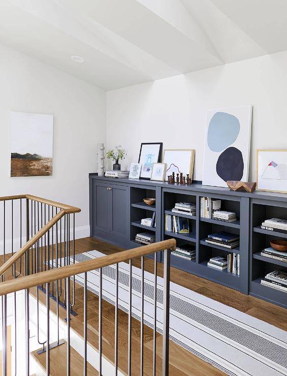 Upstairs hallway is accented with a gray and black stripe runner placed on an wood floor in front of blue built-in cabinetry finished with brass knobs and topped with framed art, as wrought iron staircase spindles are complemented with a wooden banister.