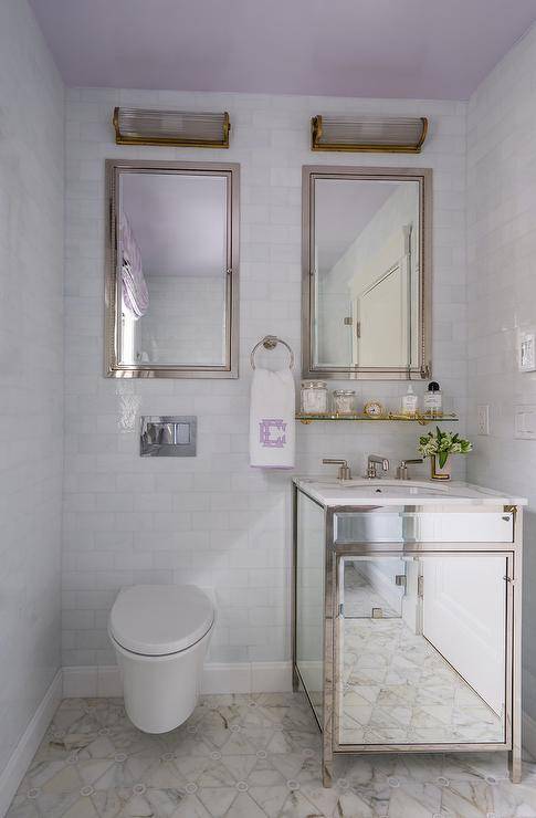 Lovely girls' bathroom features a small mirrored washstand boasting a round sink with a polished nickel faucet fixed to a marble countertop. The washstand sits on marble triangle floor tiles beneath a bras and glass vintage shelf mounted to white glass brick wall tiles beneath a polished nickel inset framed medicine cabinet. An additional medicine cabinet is fixed over a floating toilet, and both cabinets are illuminated by glass and brass sconces.