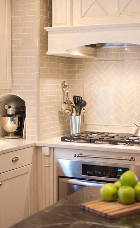 Exquisite kitchen with creamy white cabinets with white marble countertop and gray subway tile backsplash. Kitchen features paneled kitchen hood with x paneling over gray subway herringbone tile cooktop backsplash over gas range. Kitchen with small appliance nook to hold KitchenAid Mixer.