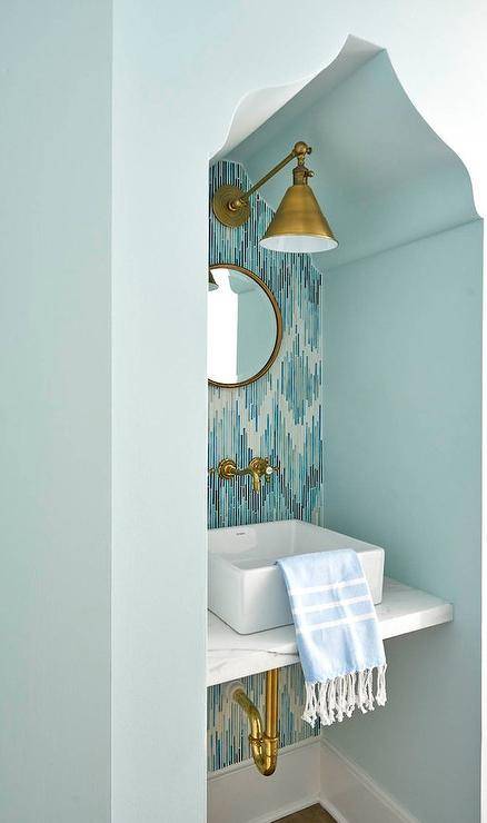A Moroccan silhouette bathroom nook boasts a honed marble sink vanity fitted with a square vessel sink mounted under an antique brass vintage style faucet mounted to blue and gray mosaic wall tiles. Above the vanity, a small round brass mirror is illuminated by a Boston Functional Library Light.