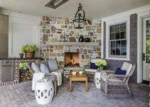 Beautifully designed covered patio features a corner rustic stone fireplace lit by carriage lantern hung over a teak slatted coffee table. The coffee table is flanked by a brown wicker sofa and a brown wicker chair, placed on gray cobblestones and matched with white rope stools.
