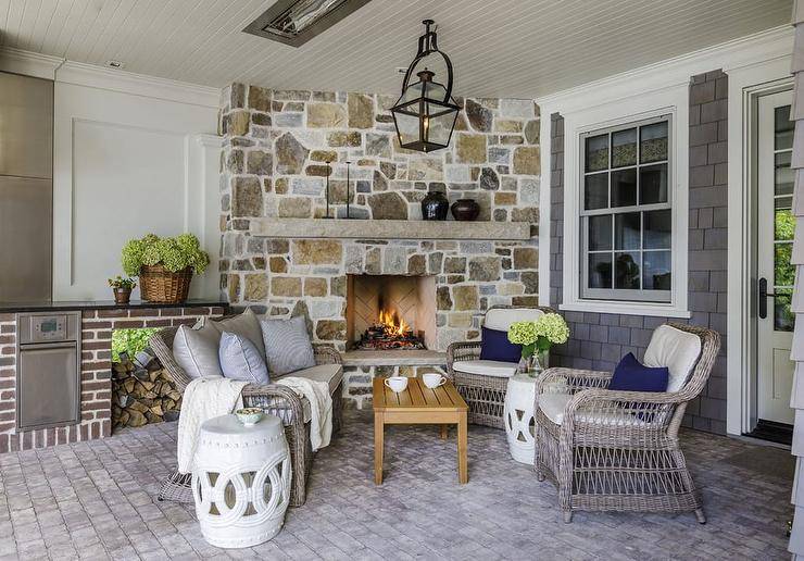 Beautifully designed covered patio features a corner rustic stone fireplace lit by carriage lantern hung over a teak slatted coffee table. The coffee table is flanked by a brown wicker sofa and a brown wicker chair, placed on gray cobblestones and matched with white rope stools.