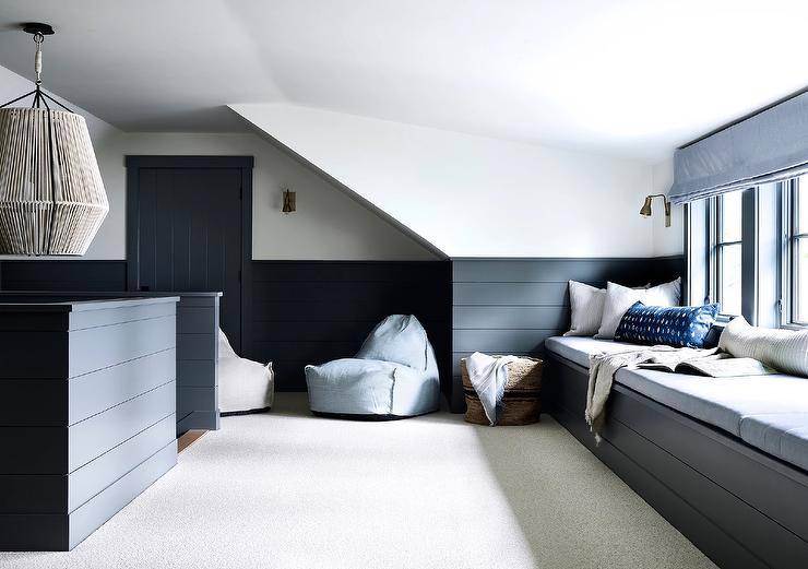 White, black, and blue attic playroom boasts a black shiplap built-in window seat topped with light gray cushions and pillows placed beneath a window dressed in a light blue roman shade. The roman shade matches blue bean bags placed in front of a wall half clad in black shiplap trim.