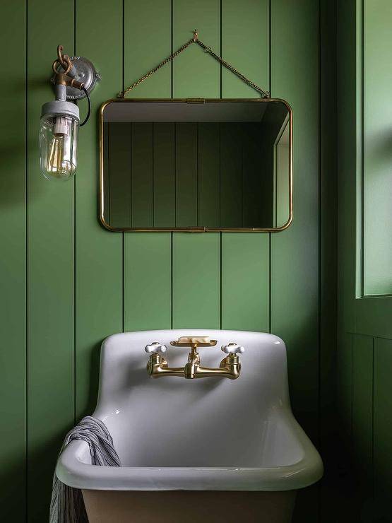 A vintage style sconce is mounted to a Kelly green shiplap wall beside a brass chain mirror hung over a vintage sink with a brass vintage style faucet.