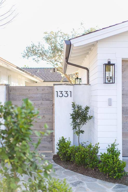 Exterior home features modern steel house numbers displayed on a white finished wooden house gate. Winding stone pavers sets a surface toward the wooden gate into a finished backyard.