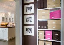 Multi functional hallway leading to kitchen features built-in shelving lined with brown, beige and hot pink storage boxes alongside dark brown walls filled with framed black and white photography over brown tiled floors.