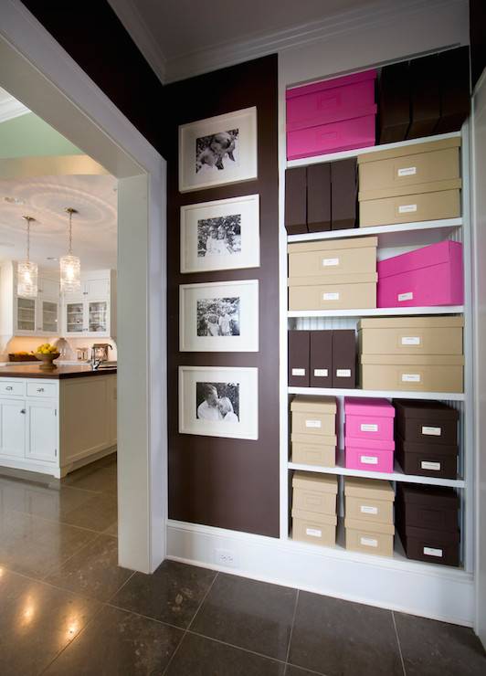 Multi functional hallway leading to kitchen features built-in shelving lined with brown, beige and hot pink storage boxes alongside dark brown walls filled with framed black and white photography over brown tiled floors.