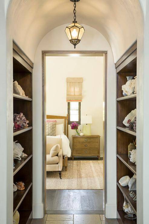 This well appointed hallway features an antique pendant hung from a barrel ceiling between facing built in wood shelves. The hallway leads to a bedroom fitted with an oak bed dressed in white hotel bedding and placed on a cream rug beside a Restoration Hardware Empire Rosette Closed Nightstand lit by a pale yellow placed beside a window covered in a beige roman shade as a cream settee sits in front of the bed.