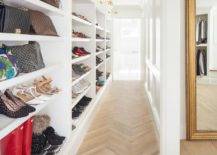Custom walk-in closet shelves organize shoes and accessories into a perfect display. Brass picture lights mount at the top of the white built-in shelves over the show storage and hallway finished with light wood herringbone pattern floors.