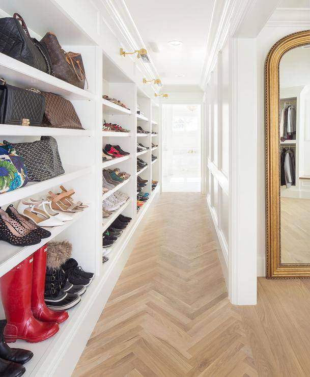 Custom walk-in closet shelves organize shoes and accessories into a perfect display. Brass picture lights mount at the top of the white built-in shelves over the show storage and hallway finished with light wood herringbone pattern floors.