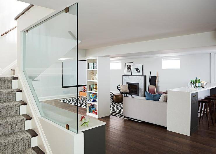 contemporary basement features a glass staircase wall positioned behind gray cabinets finished with a white countertop. The basement is also fitted with a lounge boasting a black and white and seating backless wood barstools behind a light gray sofa. The sofa is located on a black and white striped rug facing two black accent chairs complemented with gray pillows and positioned flanking a gold stool. The room is completed with a white bookcase located opposite a marble fireplace accented with a black trim and topped with framed black and white art placed between windows.