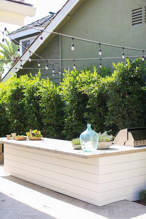 A white shiplap outdoor kitchen is accented with a concrete countertop illuminated by string lights.