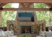 Stunning covered patio with a truss ceiling features a TV mounted on a stone fireplace, brown wicker outdoor sofas, a slatted teak coffee table and green rope accent tables.