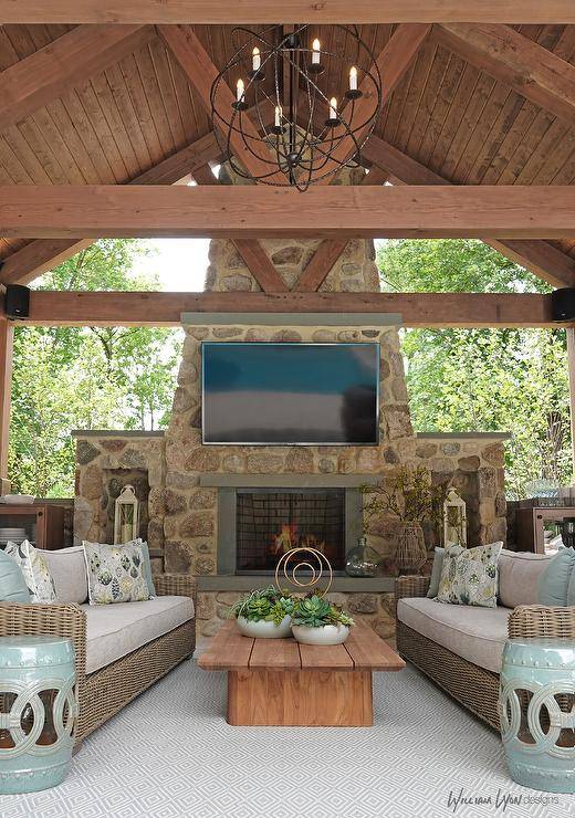 Stunning covered patio with a truss ceiling features a TV mounted on a stone fireplace, brown wicker outdoor sofas, a slatted teak coffee table and green rope accent tables.