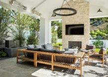 Wonderfully styled covered patio boasts a teak sectional accented with charcoal gray cushions and paired with matching teak chairs. A TV niche is located above a fireplace in a cream stone fireplace wall.