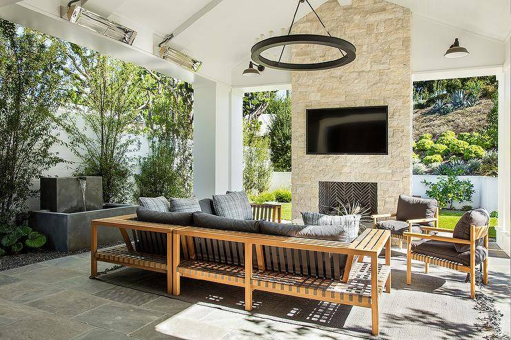 Wonderfully styled covered patio boasts a teak sectional accented with charcoal gray cushions and paired with matching teak chairs. A TV niche is located above a fireplace in a cream stone fireplace wall.