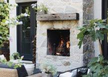 Stunning outdoor patio designed with a gray stone fireplace and a chunky reclaimed wood mantel. An outdoor wicker sofa with black rattan chairs style the space with a white geometric accent table. Black French doors bring a modern farmhouse touch to the exterior home flanking the tall stone fireplace.