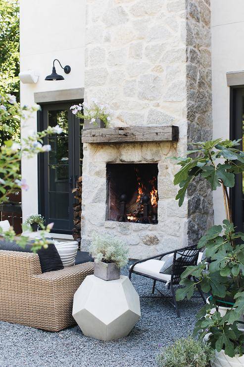 Stunning outdoor patio designed with a gray stone fireplace and a chunky reclaimed wood mantel. An outdoor wicker sofa with black rattan chairs style the space with a white geometric accent table. Black French doors bring a modern farmhouse touch to the exterior home flanking the tall stone fireplace.